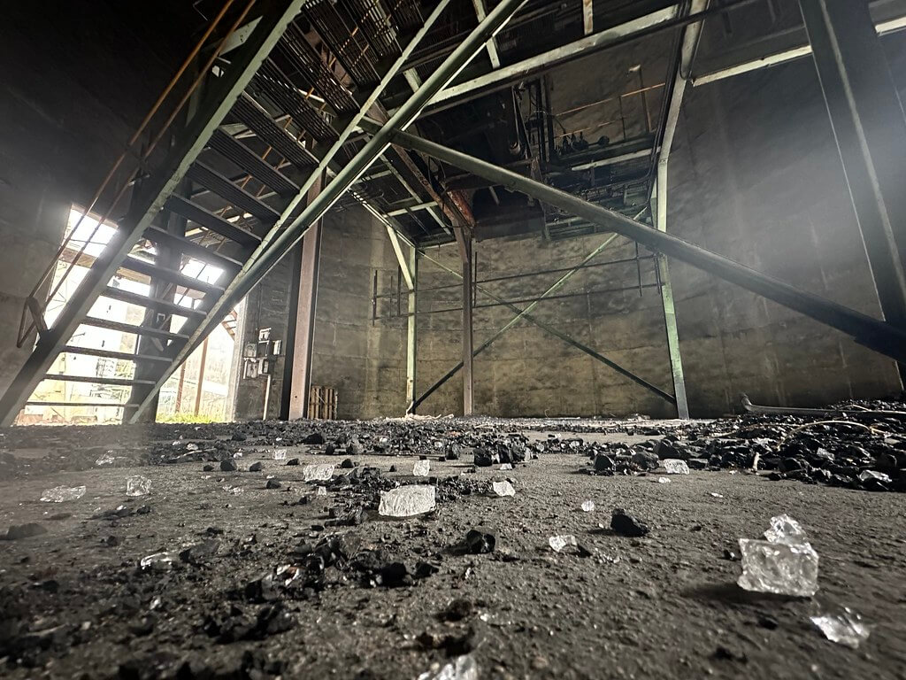 Abandoned coal prep-plant interior with steel stairs and beams; floor scattered with coal and broken glass.