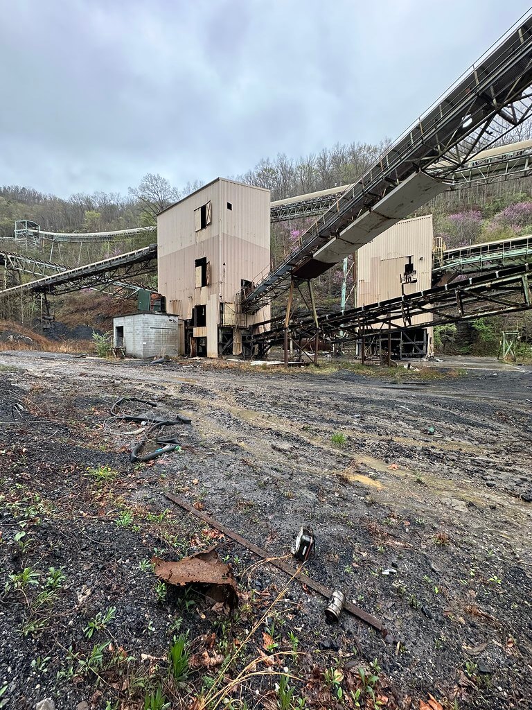 Conveyor-linked processing towers of a derelict coal plant; muddy yard littered with debris under an overcast sky.