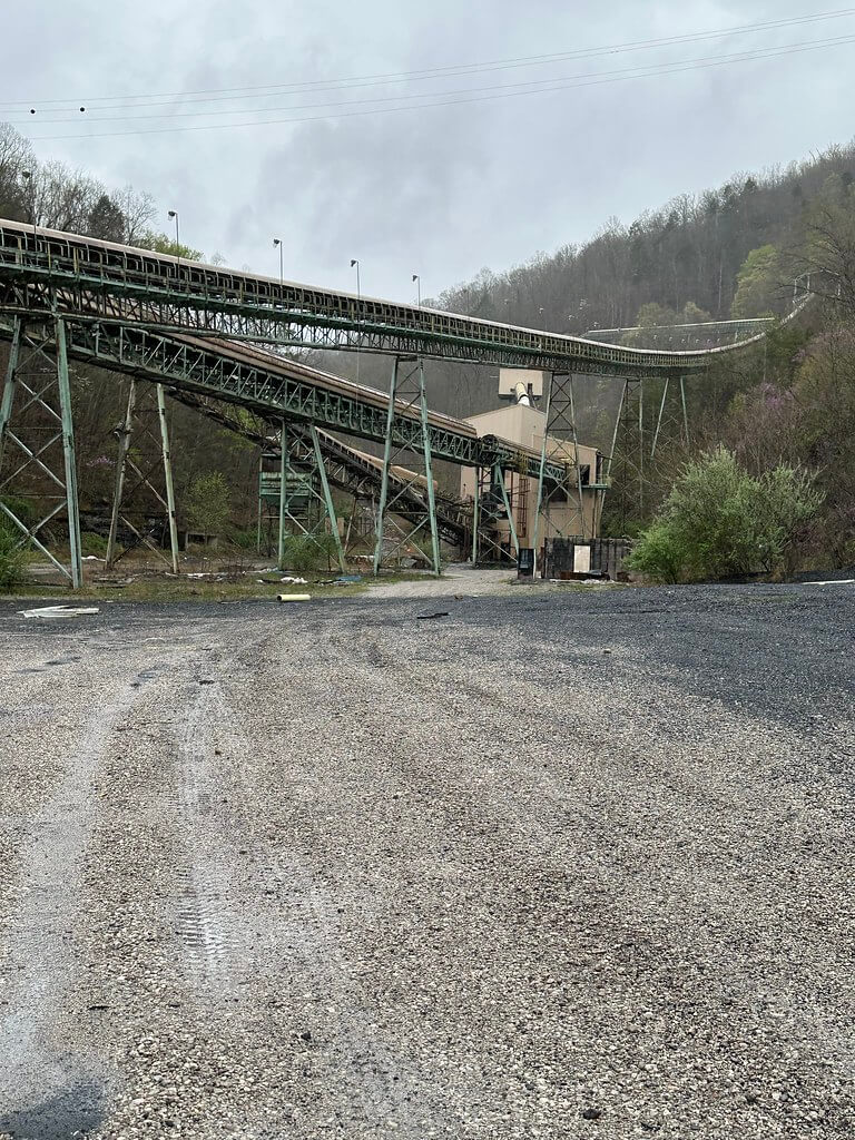 Elevated conveyors climbing a wooded hillside at a deserted coal facility; wet gravel lot in the foreground.