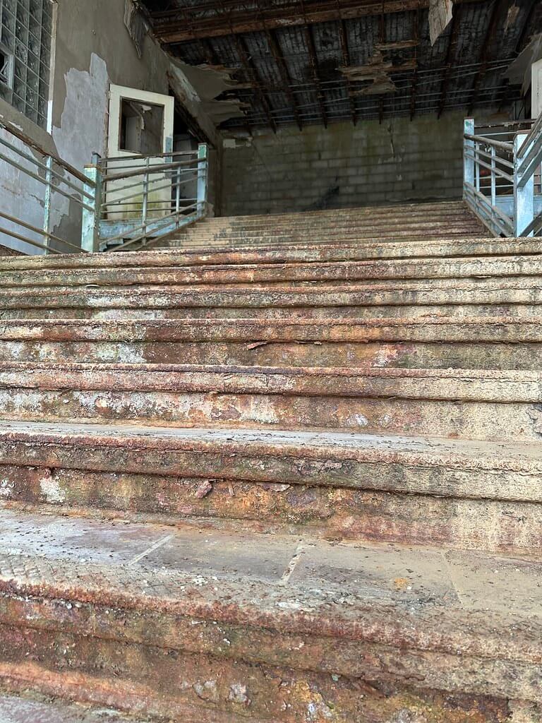 Wide, crumbling concrete steps in an abandoned school entry, with rust-streaked rails and a ceiling of exposed, damaged metal.