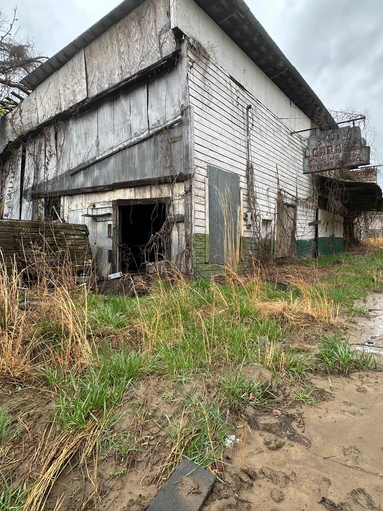 Corner view of the derelict Smith Garage, open dark bay, corrugated panels, muddy weedy lot, and a faded roadside sign under a gray sky.