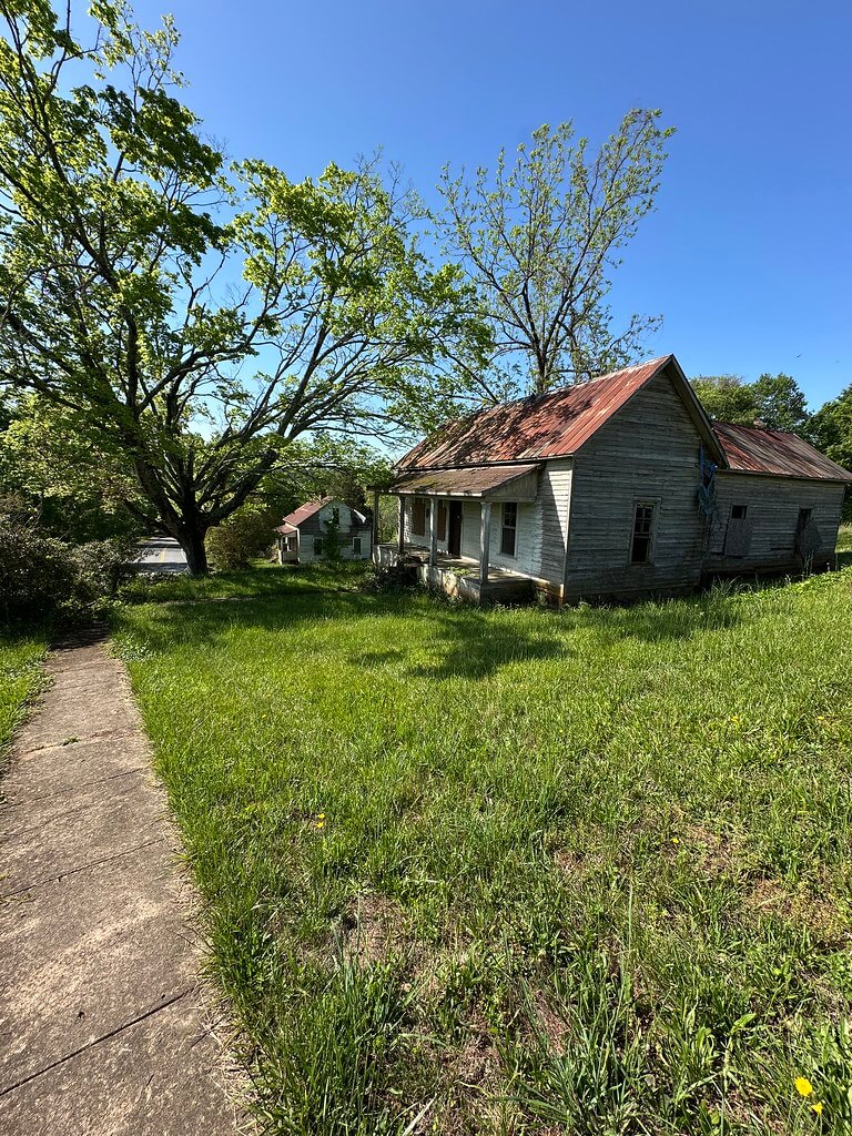 Weathered mill worker’s cottage at Henry River Mill Village with rusted metal roof and front porch, set on a grassy hill beneath a large shade tree.