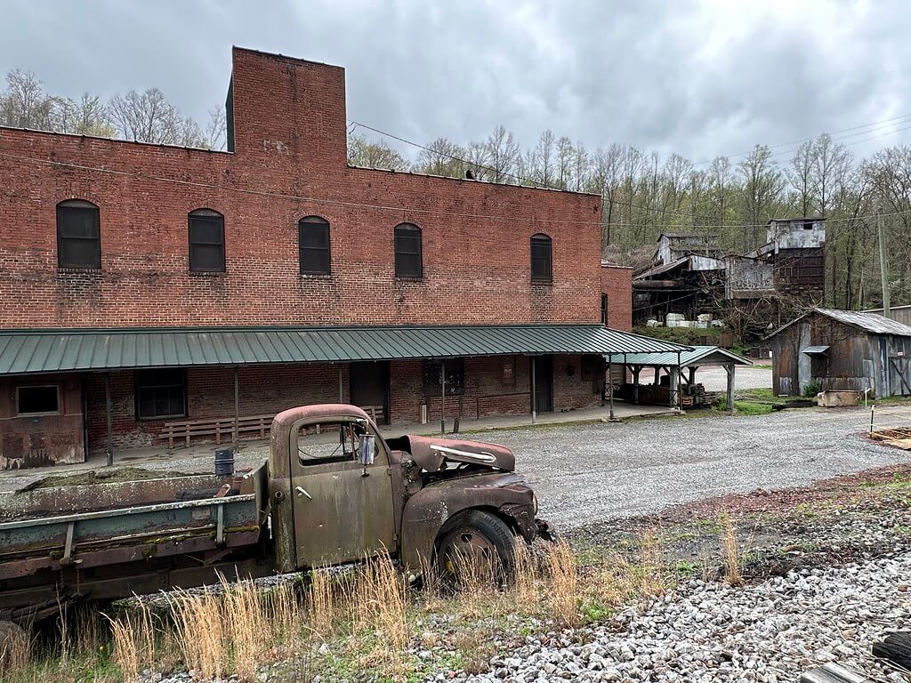 Abandoned flatbed truck in front of a two-story brick company store/warehouse with a green awning; weathered tipple structures on the hillside behind in Kitts, Kentucky.