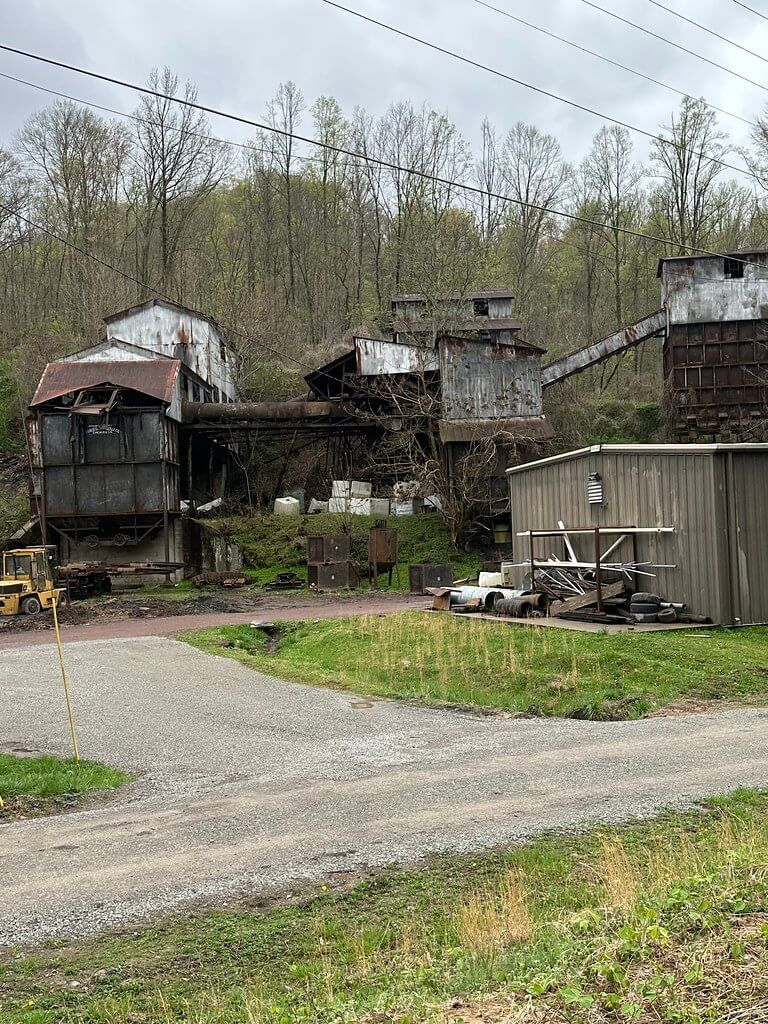 Rusted coal-tipple complex of conveyors and bins climbing a wooded slope; small metal shed, barrels, and a forklift beside a gravel road near Kitts Creek on the Clover Fork.