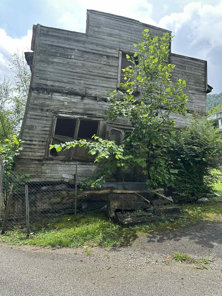Leaning false-front wooden building in Wheelwright with broken windows, chain-link fence, and saplings growing through the steps.