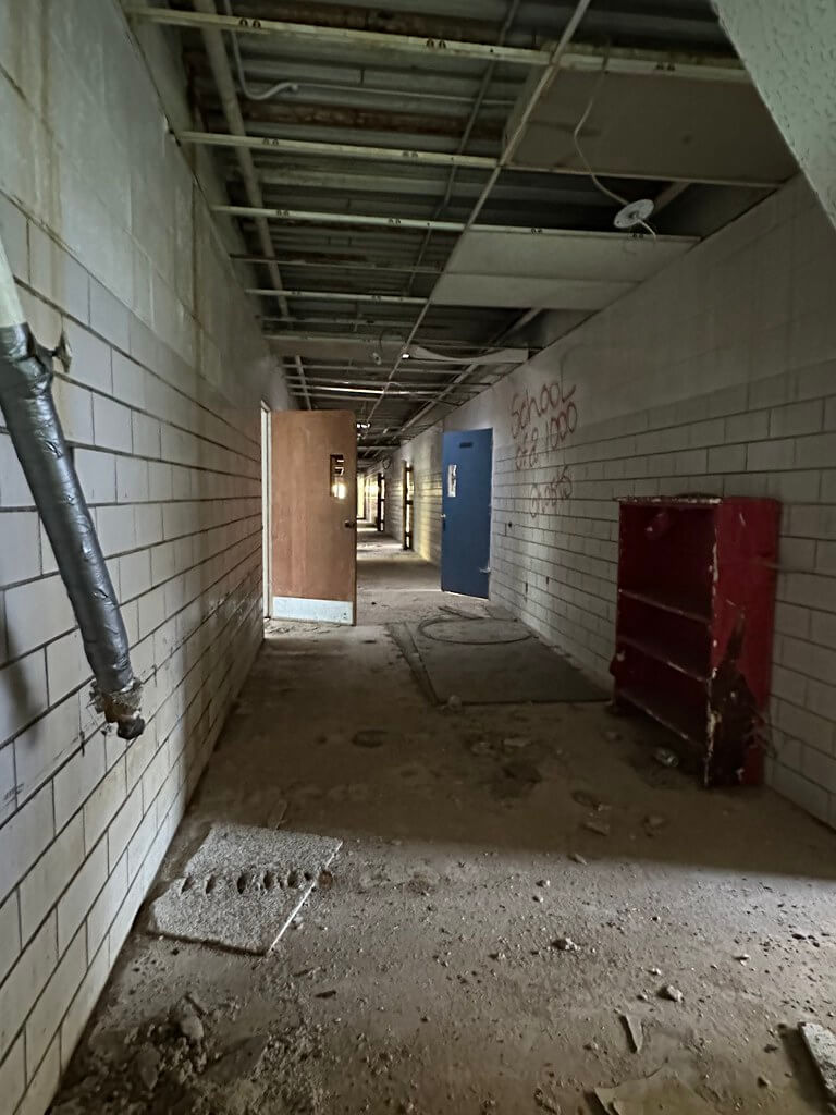 Abandoned hallway inside Cubbage Elementary: exposed ceiling grid, cinderblock walls, open doors, red bookcase, and debris underfoot.