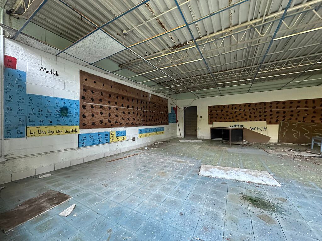 Derelict classroom at Cubbage Elementary: periodic table mural, pegboard walls, exposed metal ceiling, and a littered blue-tile floor.