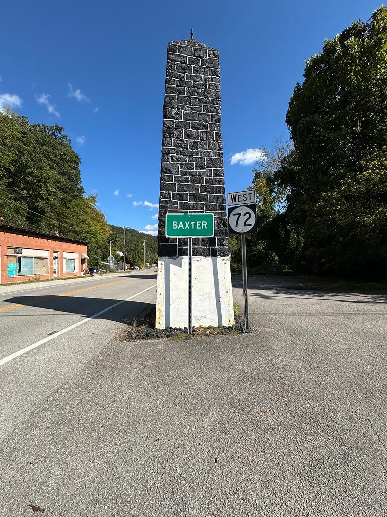 Baxter Coal Monument, a coal-block obelisk on a white base, beside a KY 72 West shield and green Baxter sign; storefronts behind.