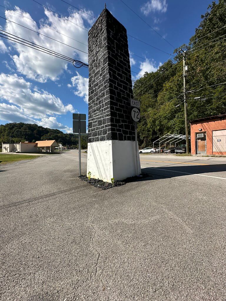 Angled view of the Baxter Coal Monument from the east side of KY 72, showing the obelisk, East 72 sign, utility lines, and nearby shops.