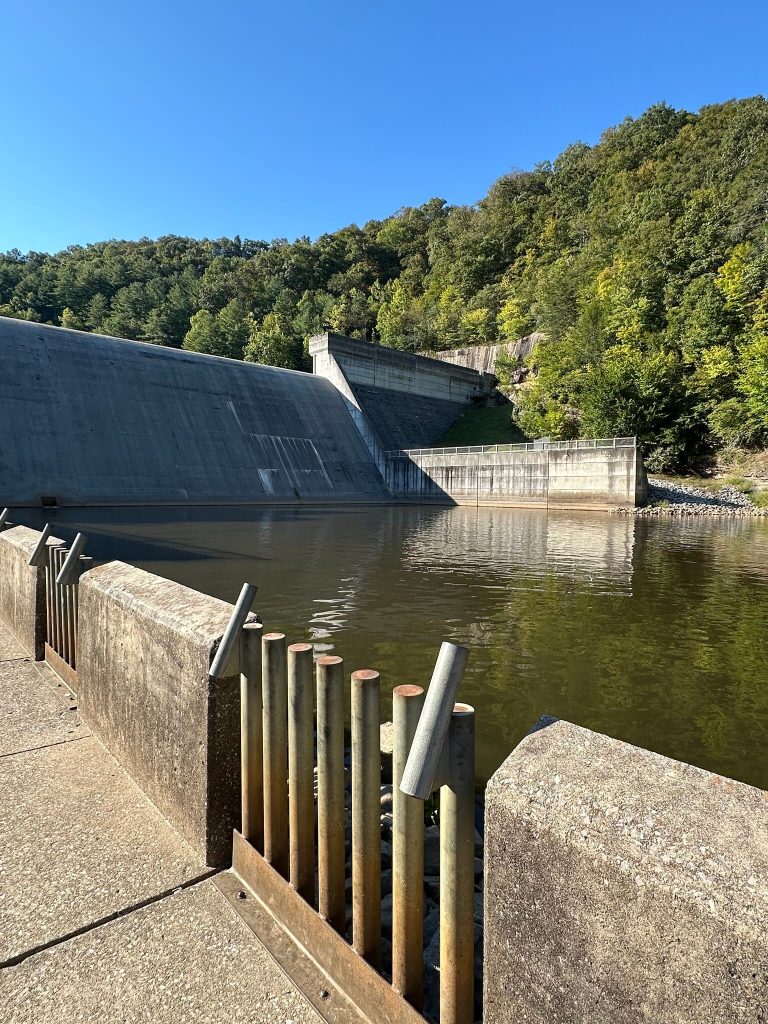 Concrete face and open spillway of Martins Fork Dam seen from the downstream walkway, with calm pool and forested ridge behind.
