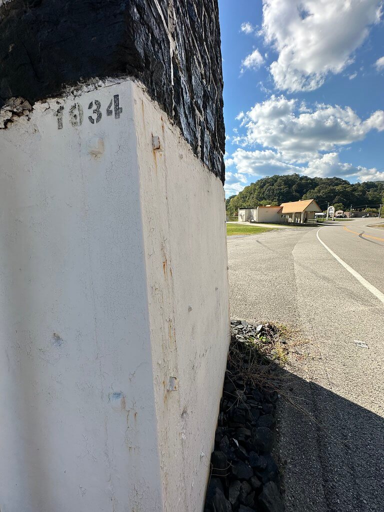 Close view of the monument’s white base with the stenciled date 1934 and anchor bolts, coal pieces at the footing, KY 72 curving in background.