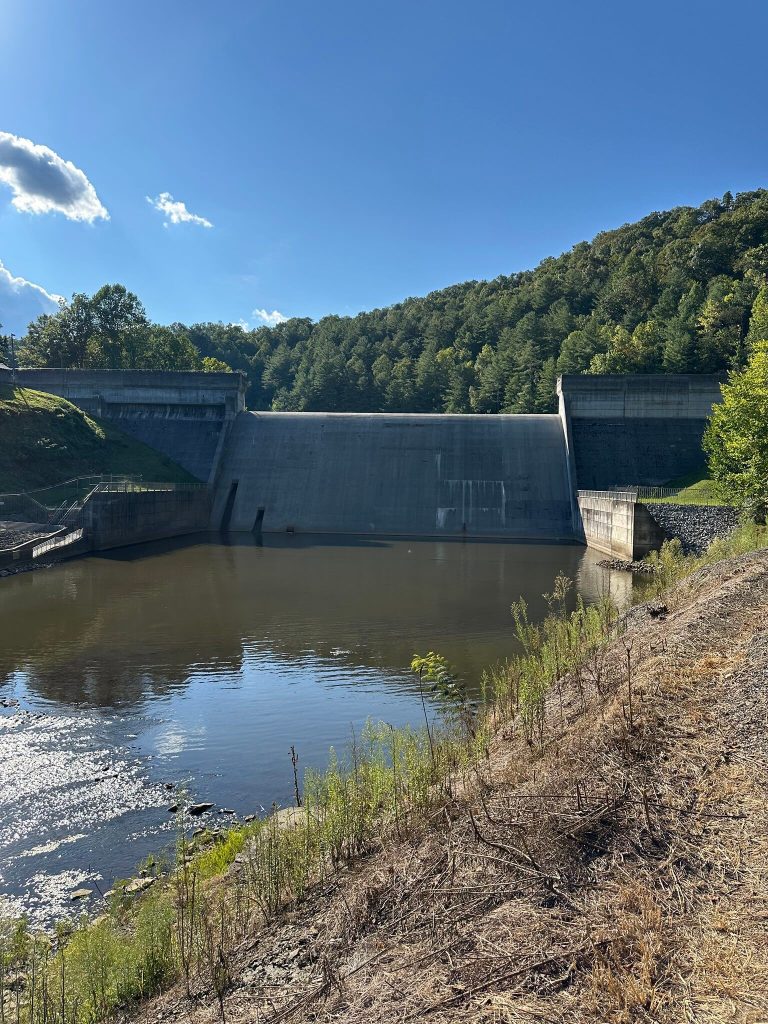 Wide view of Martins Fork Dam rising above the tailwater pool, surrounded by tree covered slopes and bright blue sky.