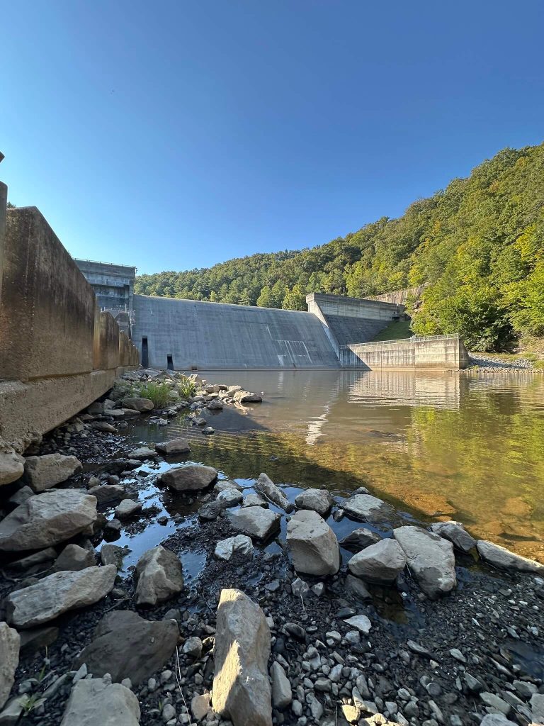 Low angle from the rocky streambank below Martins Fork Dam, looking across shallow water toward the concrete spillway and wooded hillside.