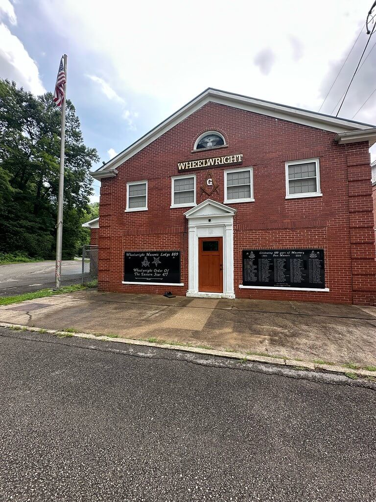 Front of the Wheelwright Masonic Lodge building in Wheelwright, Kentucky, red-brick facade with pedimented entry, arched fanlight, gold “WHEELWRIGHT” sign, Masonic emblem, memorial plaques, and a flagpole at left.