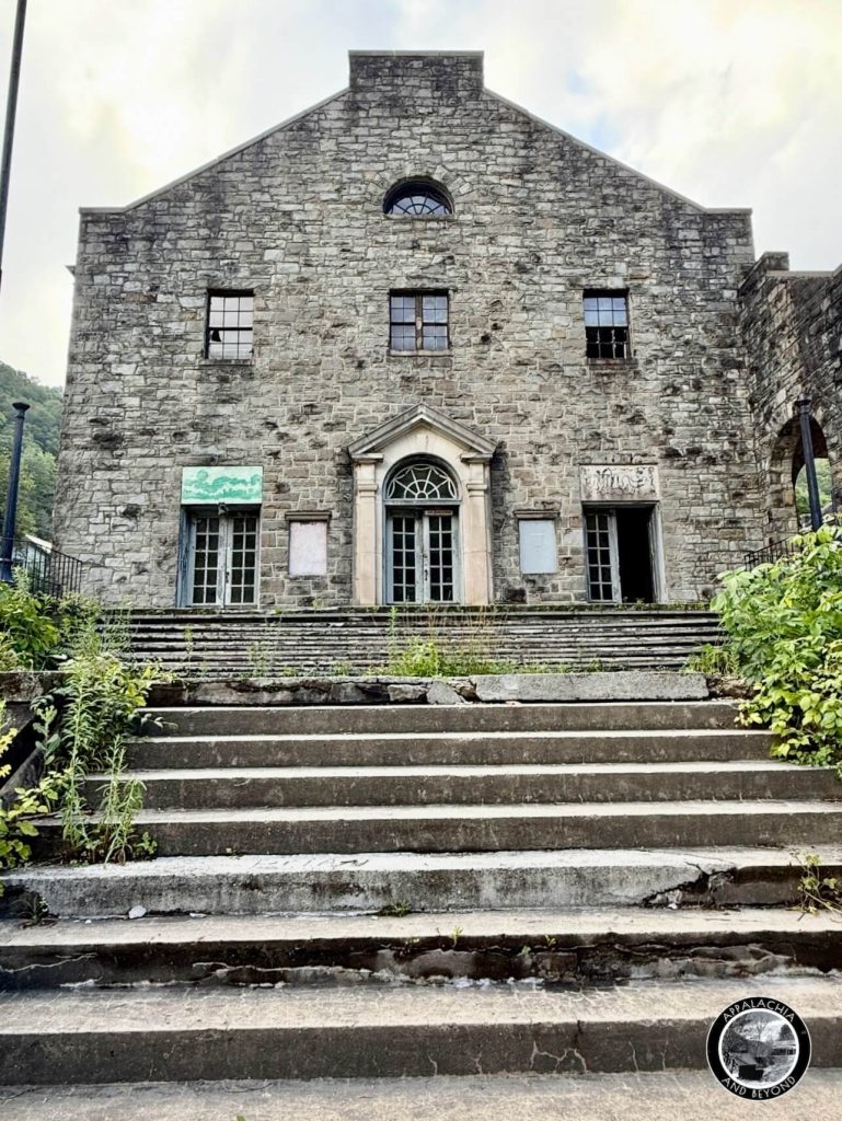 Front facade of the Itmann Company Store and Office with stone gable, arched doorway, boarded windows, and overgrown steps.