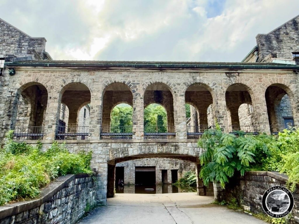 Elevated stone arcade of the Itmann complex spanning a driveway, seven round arches framing trees and hillside beyond.