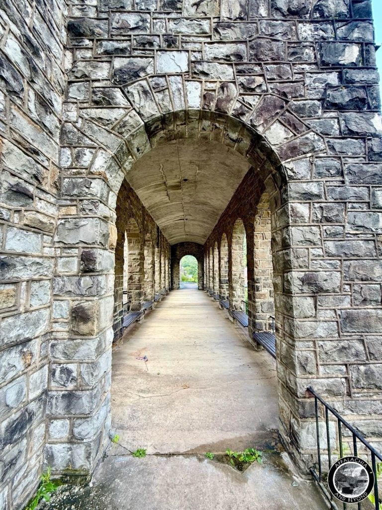 Long stone loggia at the Itmann Company Store, repeating arches and benches along a cracked concrete walkway toward daylight.