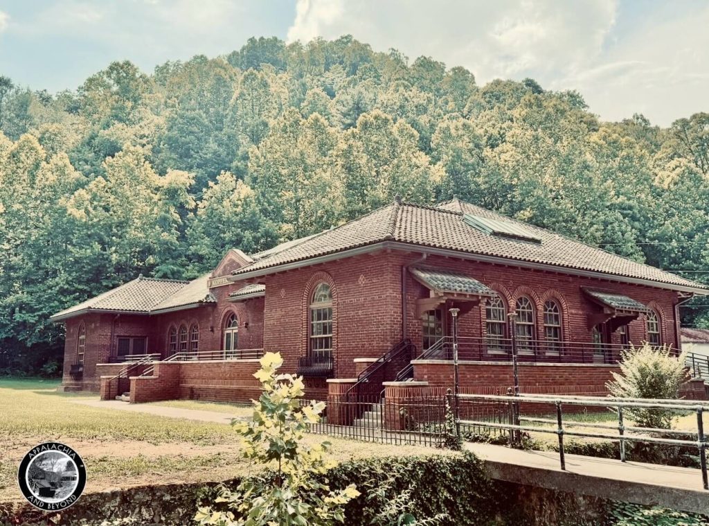Angled view of the Houston Coal Company Store beside the creek and footbridge, showing arched windows, tile roof, and skylight in Kimball, West Virginia.