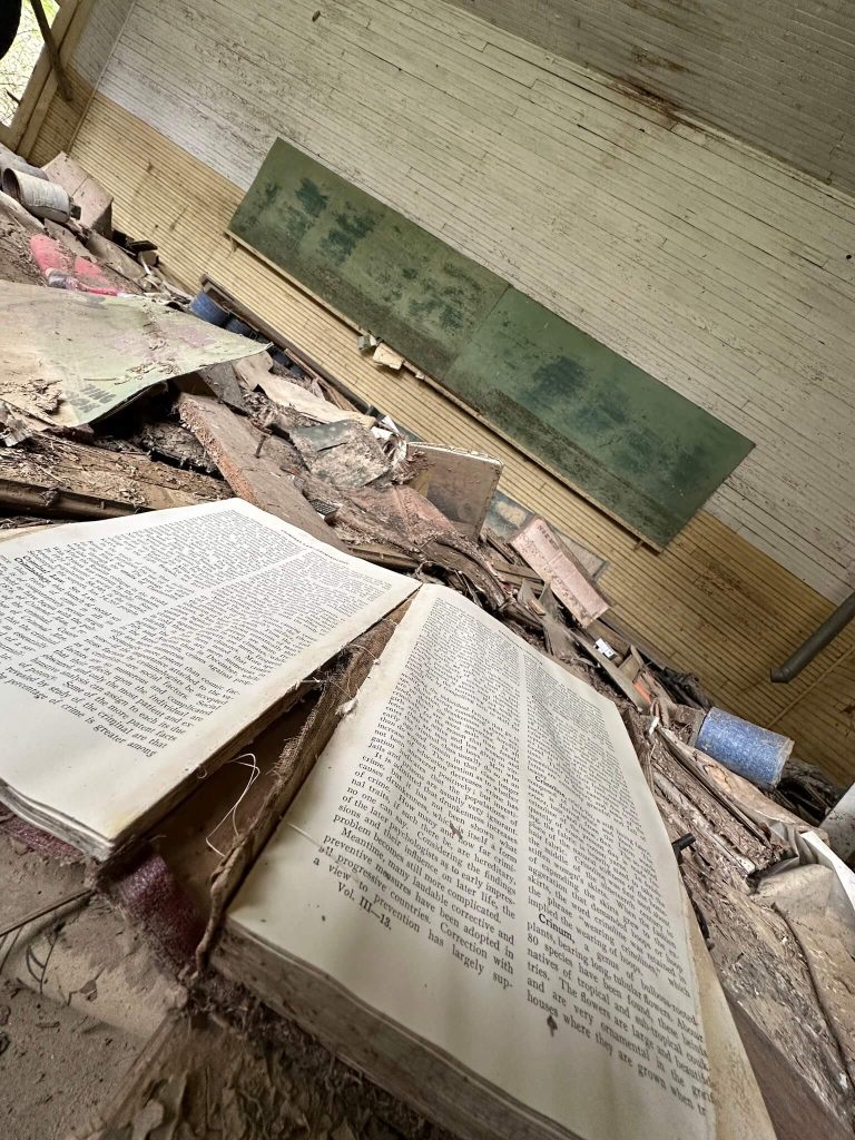 Open textbook lying amid debris inside abandoned Neely Elementary classroom in Knott County, Kentucky, with collapsed boards, peeling paint, and faded green chalkboard in background.
