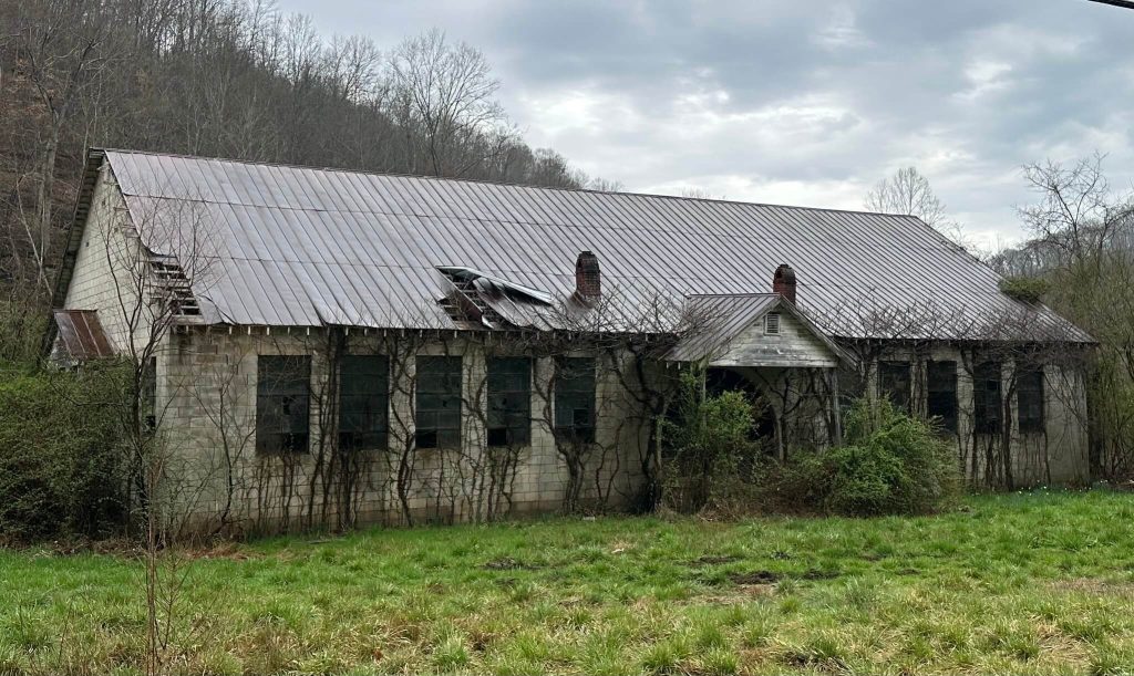 Front exterior of vacant Neely Elementary School in Knott County, Kentucky, long cinderblock building with broken windows, rusting metal roof, vines climbing walls, and overgrown grass in foreground.
