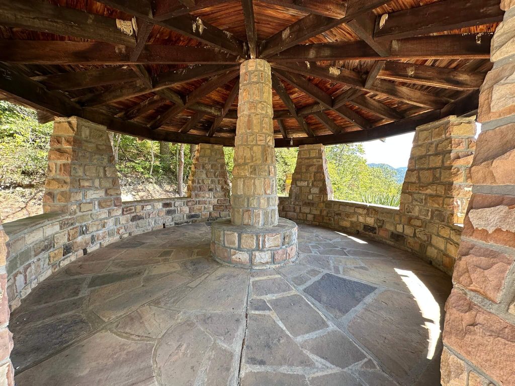 Stone pavilion at Kingdom Come State Park with a circular bench and central stone pillar under a dark wooden roof, looking out through open arches toward the surrounding forest.