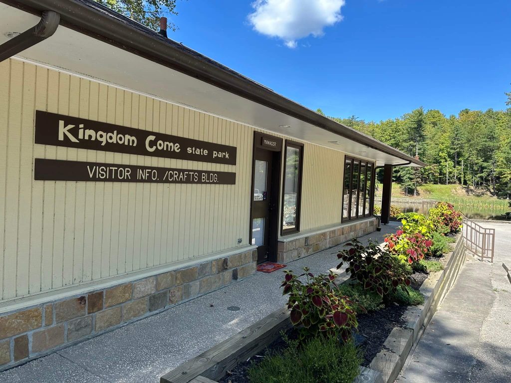 Visitor information and crafts building at Kingdom Come State Park, a long light colored structure with large windows and flower beds along the sidewalk, set beside the park lake and forest on a clear day.