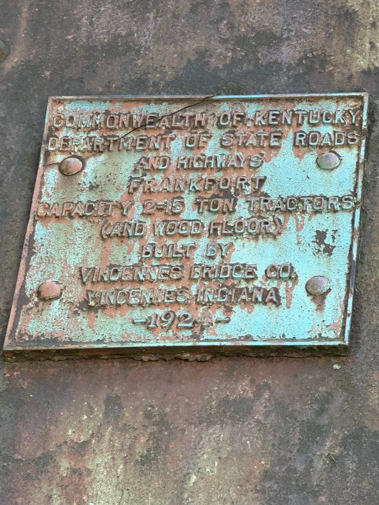 Weathered metal builder’s plaque on Baxter Bridge reading “Commonwealth of Kentucky Department of State Roads and Highways Frankfort. Capacity 2–15 ton tractors (and wood floor). Built by Vincennes Bridge Co., Vincennes, Indiana, 192–,” with green patina and rust on the riveted plate.