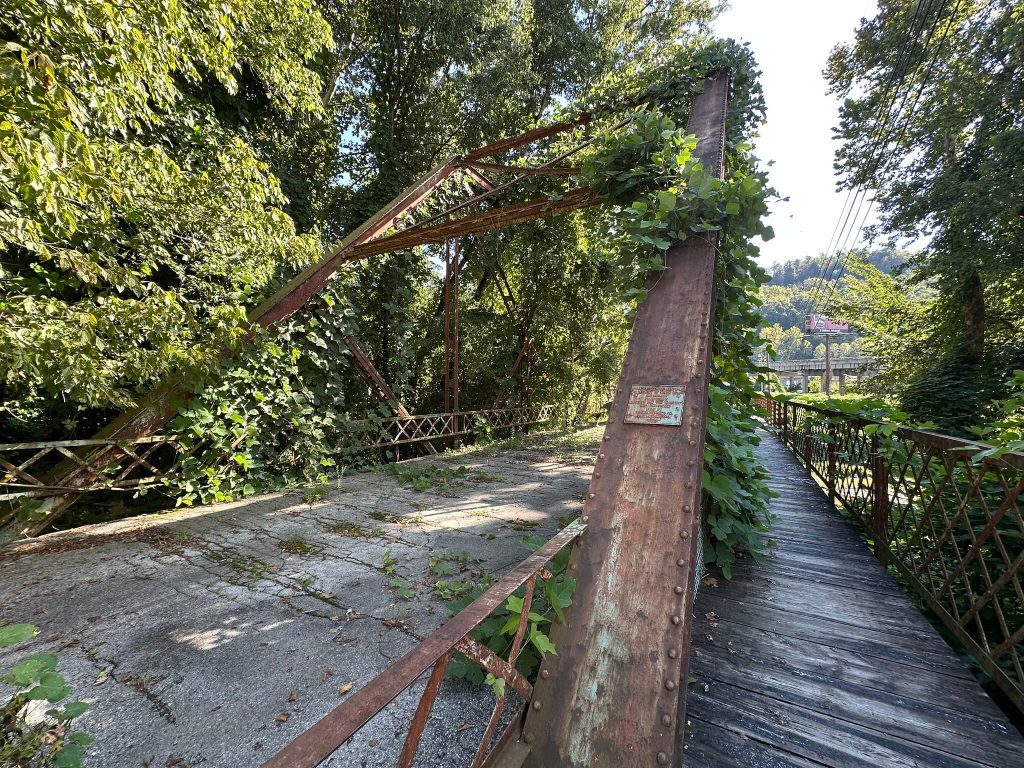 View along the abandoned Baxter Bridge in Harlan County, Kentucky, showing a rusted steel truss wrapped in vines, a cracked concrete roadway with weeds growing through it, and a narrow wooden walkway on the right, all surrounded by dense summer foliage and hills in the background.