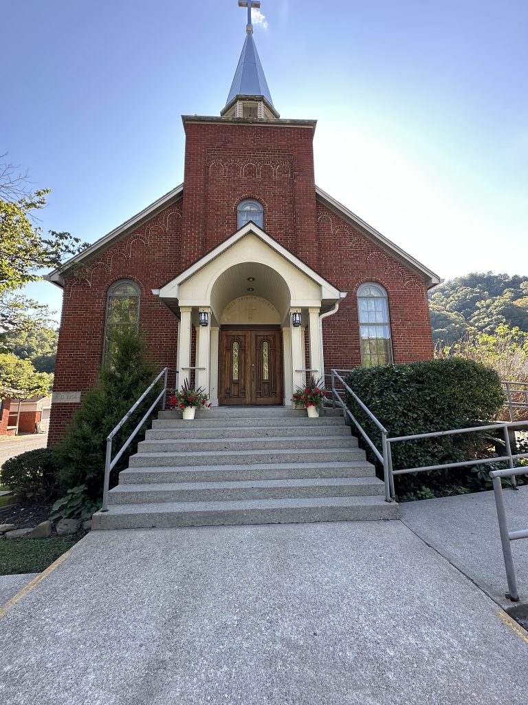 Front view of a red-brick church with a tall steeple and cross, wide concrete steps with metal railings, and flower pots on either side of the wooden double doors, set against a backdrop of green hills and blue sky.