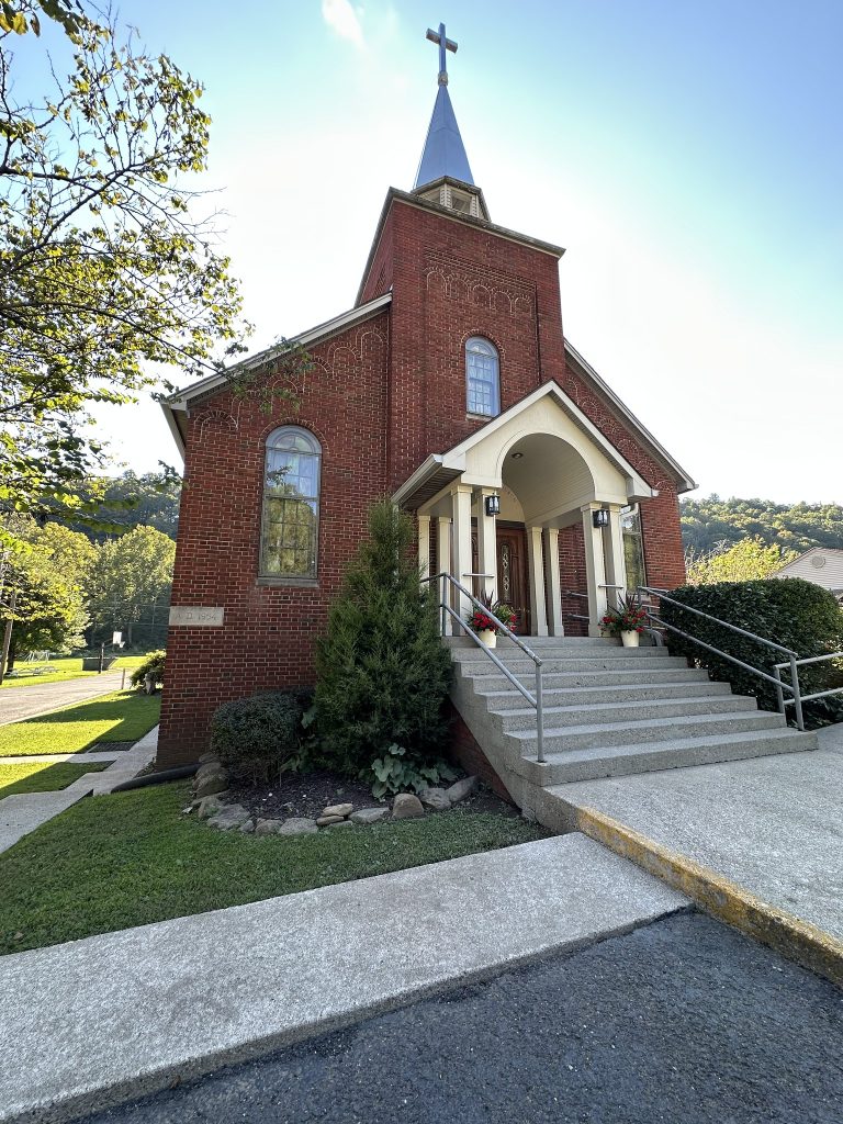 Angled side view of the same red-brick church, showing arched windows, neatly trimmed shrubs and small trees, and concrete walkways leading up to the entrance.