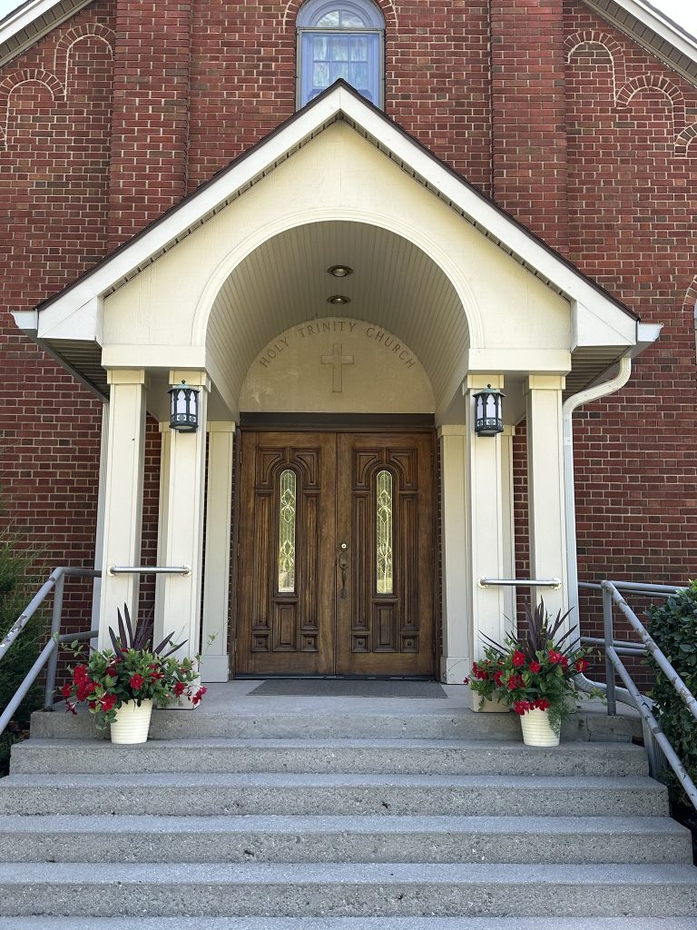 Close-up of the church entrance with wooden double doors under a small white portico, flanked by hanging lanterns and planters of red flowers, with “HOLY TRINITY CHURCH” engraved above the doorway.