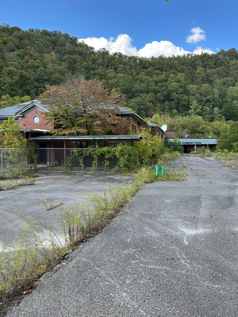 Approach to the abandoned Millard Elementary School from the driveway. A tall red brick section of the building with a circular window and sagging metal awning stands on the left behind a chain-link fence. The wide asphalt drive and parking area are cracked and filled with grass and weeds. In the distance, under a flat canopy, part of the sign reading “Elementary School” is visible, backed by a steep, tree-covered hillside and a bright blue sky with clouds.