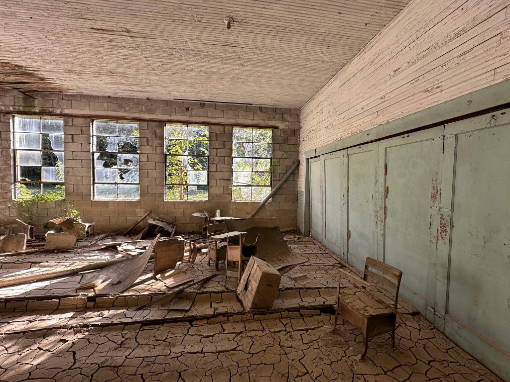 Sunlit interior of abandoned Neely Elementary classroom with cracked mud floor, overturned wooden desks, broken glass block windows, and pale green sliding doors lining one wall.