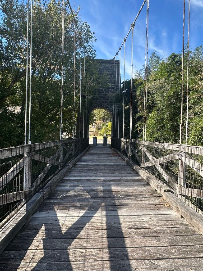 View down the wooden deck of Pauley Bridge, a narrow suspension footbridge with cables overhead, leading to a stone tower framed by trees under a blue sky.