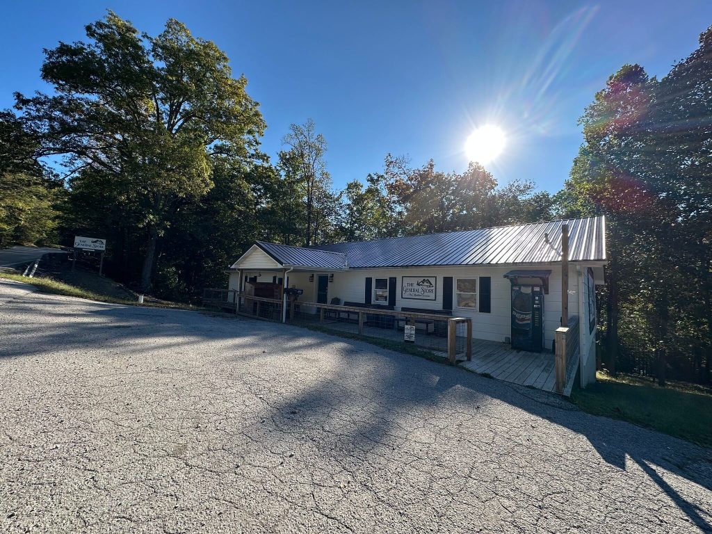 Wide view of The General Store at Pine Mountain Crossing, a long white building with a metal roof beside a mountain road, trees and bright morning sun in the background.