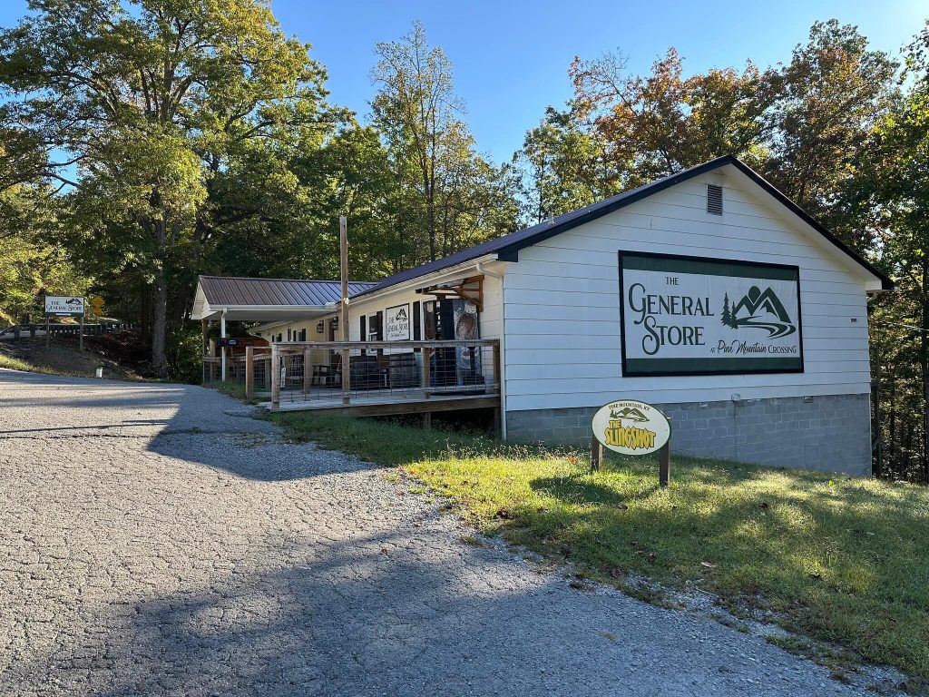 Angled view of The General Store at Pine Mountain Crossing, showing the large wall sign, front porch, and sloping paved road surrounded by trees on Pine Mountain.