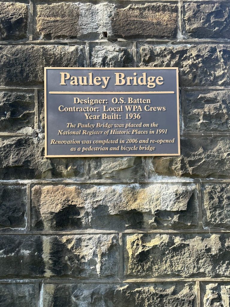 Close-up of a bronze plaque mounted on a stone tower reading “Pauley Bridge,” with details about the 1936 WPA-built bridge and its renovation as a pedestrian and bicycle bridge.
