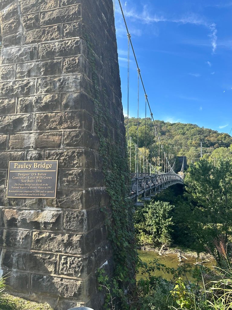Side view of Pauley Bridge showing the stone tower with plaque in the foreground, suspension cables stretching across a green river valley, and wooded hills in the background under a clear sky.
