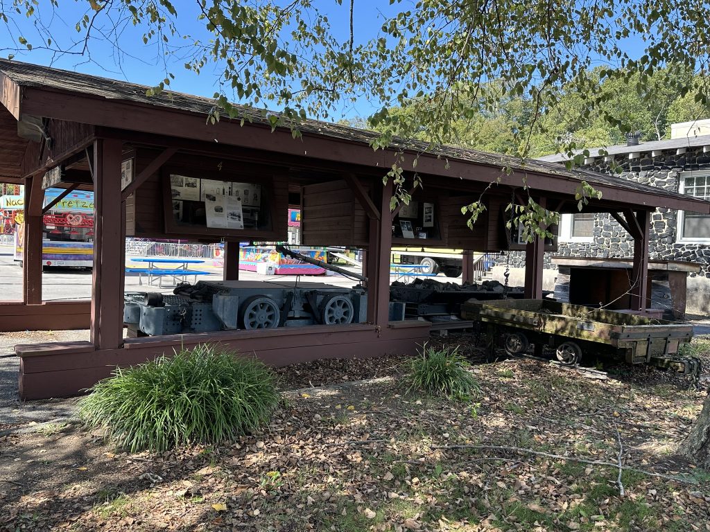 Open-sided wooden pavilion in a small park displaying historic coal-mining equipment, including gray mine carts and machinery on tracks, with fair rides and a food truck visible in the background.