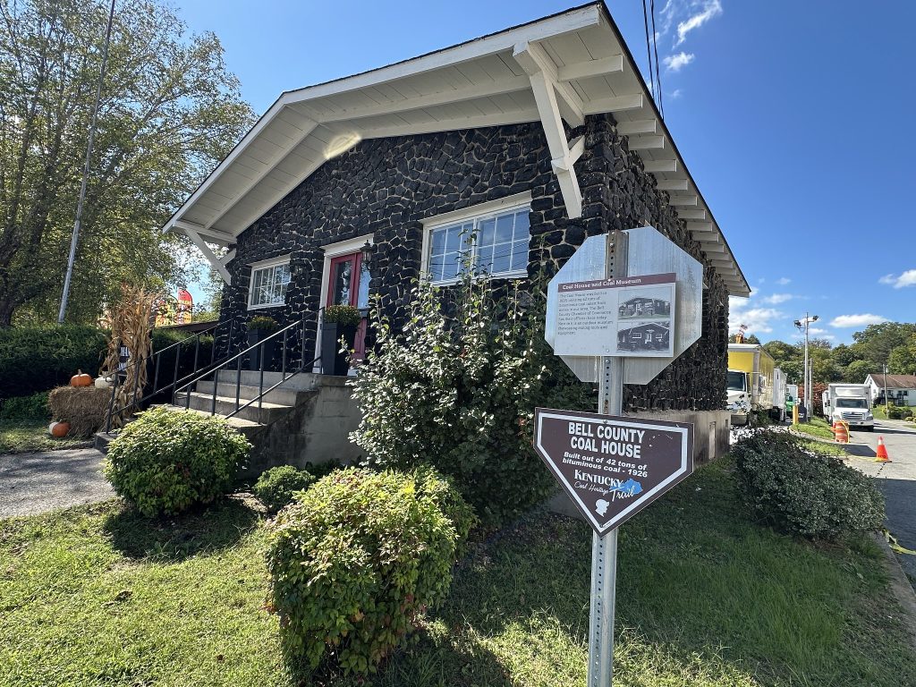 Angled view of the Bell County Coal House, showing its black coal-block exterior, red front door, surrounding shrubs and grass, and roadside signs identifying it as “Bell County Coal House” on the Kentucky Coal Heritage Trail.
