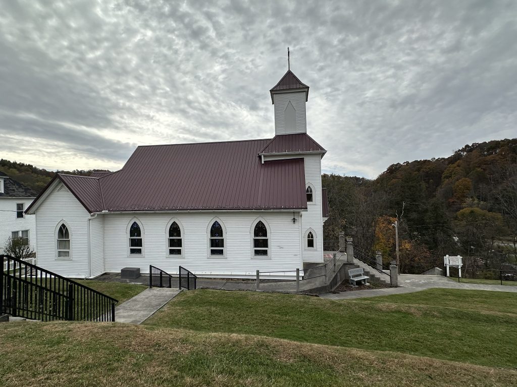 A white wooden church with a red metal roof and central steeple sits on a grassy hillside under a cloudy gray sky. The side wall shows a row of pointed-arch windows, and concrete walkways with black railings lead up to the building. Forested hills with autumn color rise in the background.