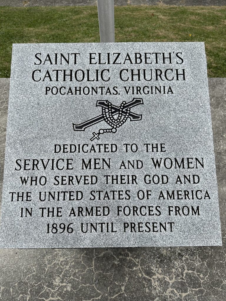 Close-up of a gray granite dedication tablet outside Saint Elizabeth’s Catholic Church in Pocahontas, Virginia. The stone is engraved with a rosary and cross and text honoring service men and women who have served God and the United States in the armed forces from 1896 to the present.