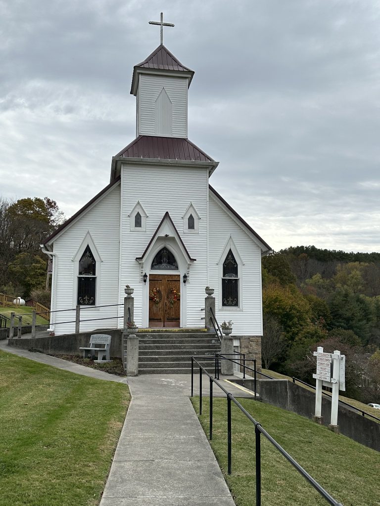Front view of Saint Elizabeth’s Catholic Church, a white clapboard building with a tall square steeple topped by a cross and a red metal roof. A straight concrete walkway with black railings leads up a gentle slope to a set of stairs and double wooden doors decorated with wreaths. Trees with fall foliage cover the hills behind the church, and a white signboard stands to the right of the path.