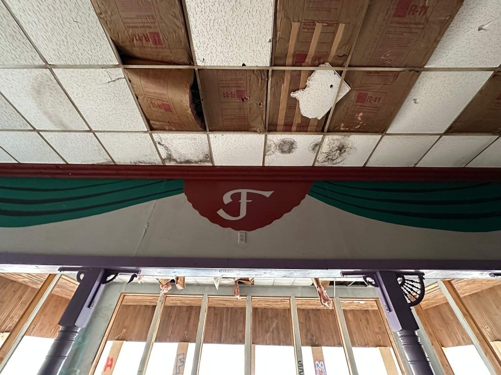 Interior of an abandoned building with stained and missing drop-ceiling tiles, exposed insulation, and a painted stage-like arch with green curtains and a red circle featuring a white letter F.