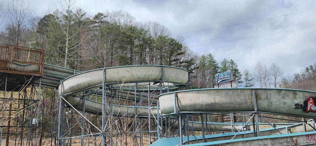 Rusting, empty water slides on metal supports at an abandoned outdoor park, with leafless trees and a cloudy sky in the background and a weathered ‘Fugates’ sign in the distance.