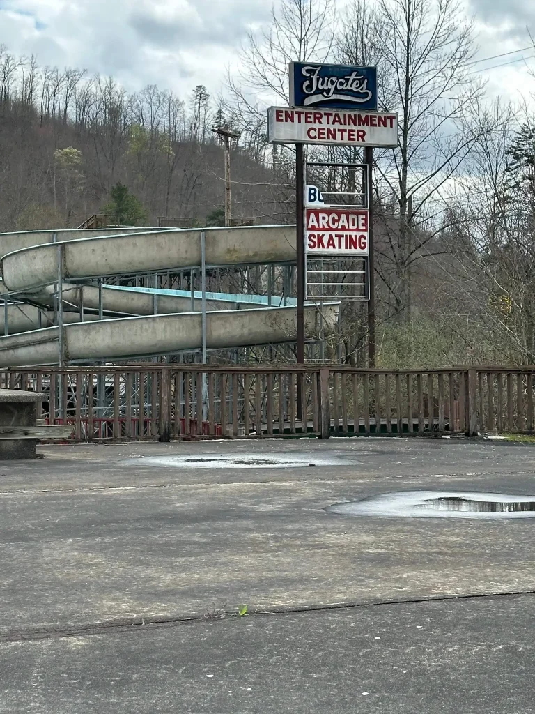 This exterior photograph shows the sign for Fugate’s Entertainment Center, advertising arcade and skating, with a large outdoor slide structure beside it. The worn pavement, empty lot, and surrounding Appalachian hillside emphasize the site’s abandoned or long-unused condition.