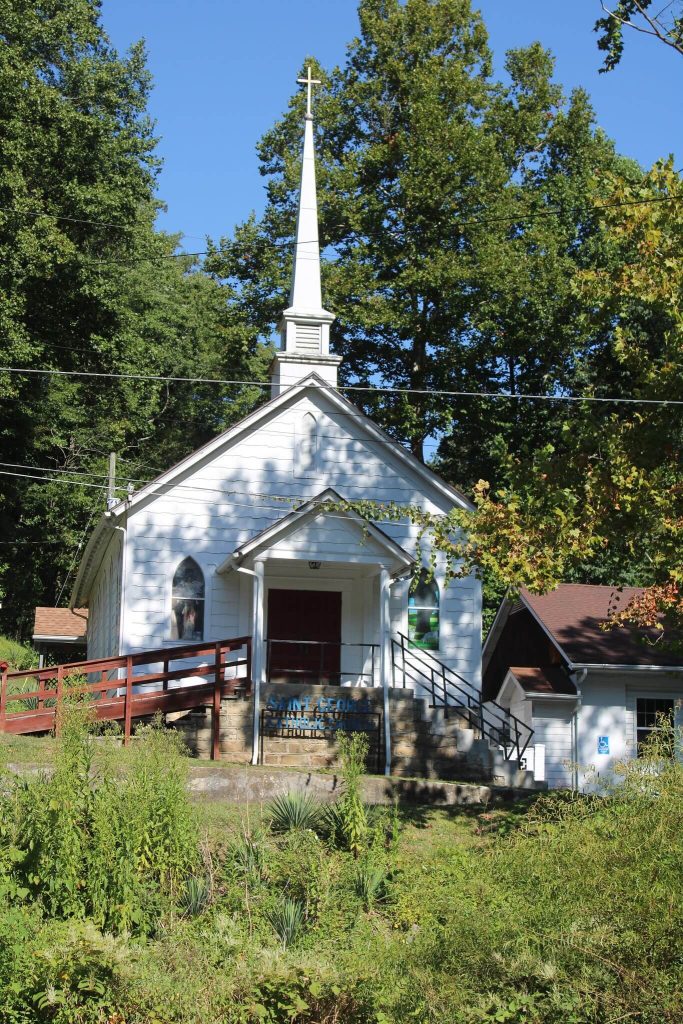 Front view of St. George Catholic Church, a small white frame church with a tall steeple and cross, steps and a ramp, framed by trees.