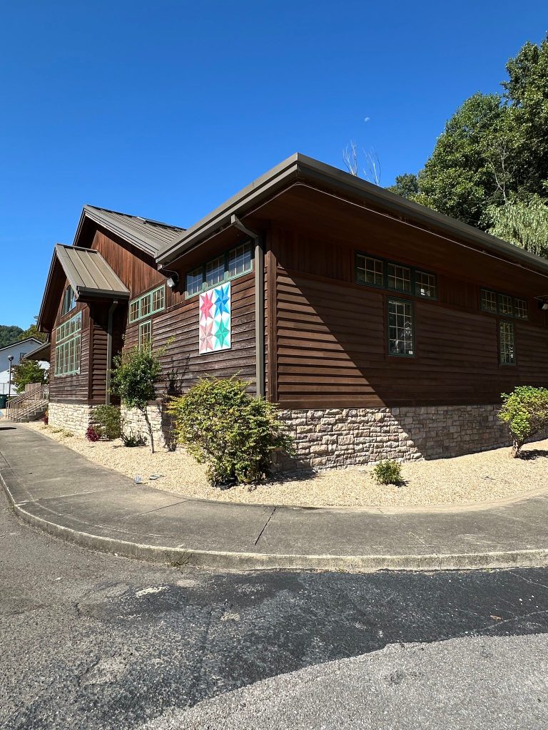 Side view of the Jenkins Public Library building in Jenkins, Kentucky, a wood-and-stone structure with a colorful quilt-block mural on the wall under a clear blue sky.