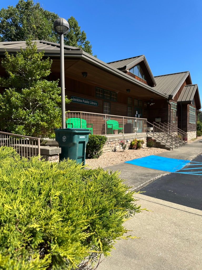 Front entrance of the Jenkins Public Library in Jenkins, Kentucky, showing the library sign, green chairs on the porch, landscaping, and accessible parking spaces.