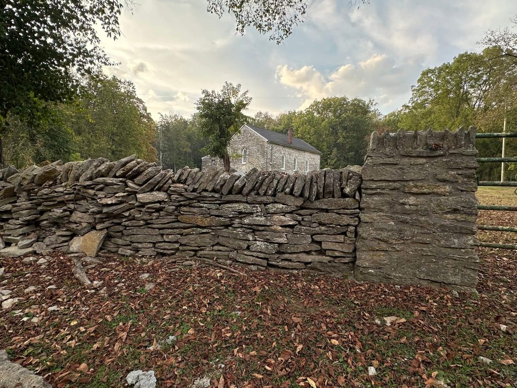 Curving dry-stacked stone wall and corner pillar in front of a small limestone church, with autumn leaves on the ground and trees in the background.